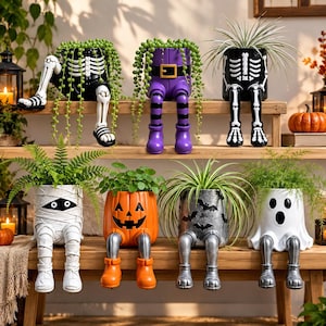 May include: A collection of seven ceramic planters shaped like Halloween characters, including skeletons, a mummy, a jack-o'-lantern, a bat-patterned pot, and a ghost, all featuring dangling legs and filled with various green houseplants.