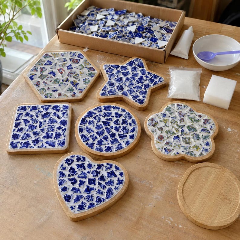May include: A mosaic craft kit featuring six wooden bases in hexagon, star, square, circle, flower, and heart shapes. Includes blue and white ceramic tile shards, a box of loose tiles, a tube of adhesive, white grout powder, and a sponge.