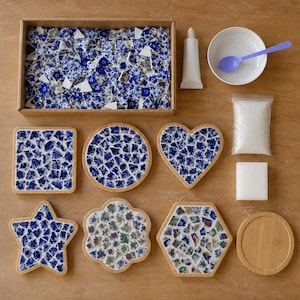 May include: A mosaic craft kit featuring a rectangular box filled with blue and white ceramic shards, a tube of adhesive, a white bowl with a purple spoon, a bag of white grout, a white sponge, and various wooden bases in square, round, heart, star, and hexagonal shapes.