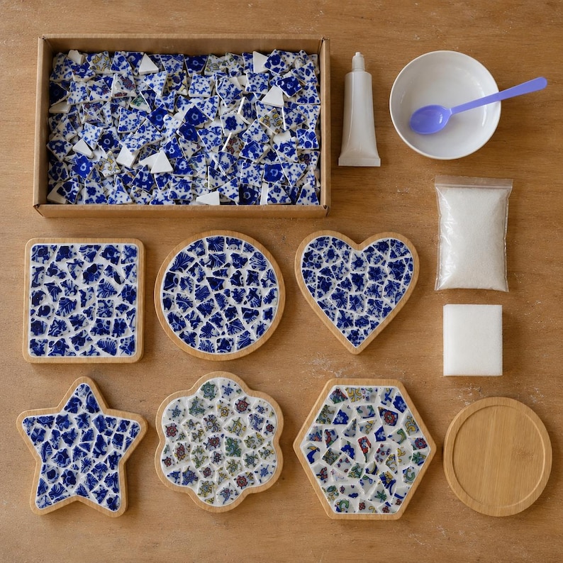 May include: A mosaic craft kit featuring a rectangular box filled with blue and white ceramic shards, a tube of adhesive, a white bowl with a purple spoon, a bag of white grout, a white sponge, and various wooden bases in square, round, heart, star, and hexagonal shapes.