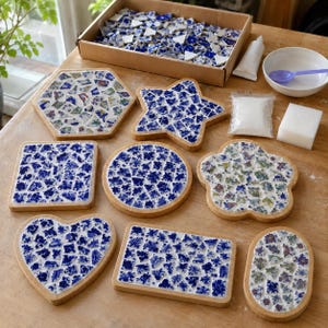 May include: A collection of eight wooden bases with blue and white mosaic tile patterns in various shapes including a hexagon, star, square, circle, flower, heart, and rectangles, alongside a box of loose tiles, adhesive, and a white sponge.