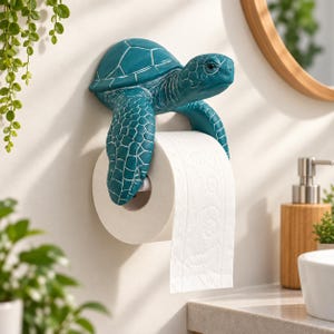 Könnte beinhalten: Ein wandmontierter Toilettenpapierhalter in Form einer blaugrünen Meeresschildkröte mit texturiertem Panzer und Flossen. Die vorderen Flossen der Schildkröte ragen nach unten und halten eine Rolle weißes Toilettenpapier auf einer Stange.