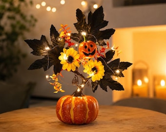 May include: A decorative tabletop centerpiece featuring a base shaped like a round orange pumpkin. A central stem supports black maple leaves, yellow sunflowers, and a small orange jack-o'-lantern, all illuminated by thin, glowing string lights.