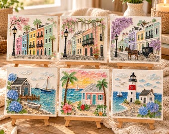 May include: Six square canvas art pieces on wooden easels featuring textured pebble and bead mosaics. Scenes include colorful townhouses, a horse-drawn carriage, a coastal cottage with a sailboat, a tropical beach house, and a lighthouse by the sea.