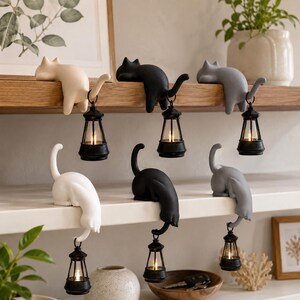 Può includere: Sei statuette a forma di gatto in bianco, nero e grigio, ognuna delle quali regge una piccola lanterna nera con una luce brillante. Tre gatti sono posizionati sul bordo di una mensola in legno, mentre altri tre siedono su una mensola bianca.