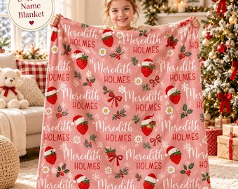 Puede incluir: Una manta de forro polar rosa con un patrón repetitivo de fresas con gorros de Papá Noel, margaritas blancas, ramas de acebo y el texto Meredith Holmes en letra cursiva blanca y fuente serif roja, con copos de nieve.