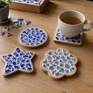 May include: A set of wooden coasters featuring mosaic tile inlays. One coaster is circular, one is star-shaped, and one is flower-shaped. The tiles display blue and white floral patterns, while a beige ceramic mug sits on a square coaster.