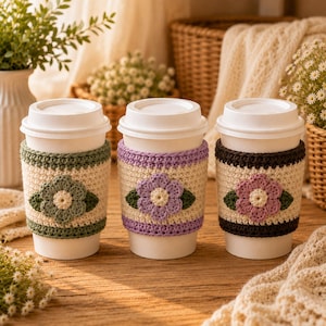 May include: Three white disposable coffee cups with removable crochet sleeves. Each sleeve features a cream-colored center with a crocheted flower in sage green, lavender, or dark pink, accented by two dark green leaves and matching colored borders.