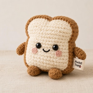 May include: A crochet plush toy shaped like a slice of bread with a cream-colored center and brown crust. It features black plastic eyes, a stitched smile, pink cheeks, small arms, and legs. A white fabric tag on the side reads Pocket Toast.
