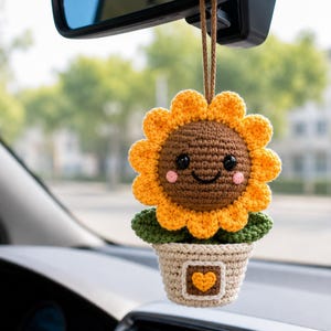 May include: A crocheted sunflower car hanging ornament featuring a brown centre with black eyes and a smile, surrounded by yellow petals. The flower sits in a beige pot with a small brown square containing a yellow heart, suspended by a brown cord.