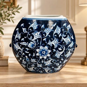 May include: A round ceramic vase featuring a deep blue glaze with intricate white floral and vine patterns. The vessel has a wide, open top and small side handles, showcasing a glossy finish and a symmetrical, detailed botanical design.