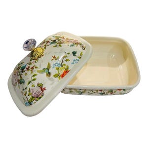 May include: A rectangular ceramic butter dish with a cream-colored interior and a lid featuring a colorful floral and hummingbird pattern. The lid is topped with a faceted, clear crystal-like knob set on a gold-toned base, adding a decorative touch.