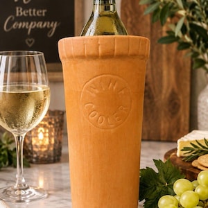 May include: A cylindrical terracotta wine cooler with a textured rim and a circular embossed stamp on the front that reads WINE COOLER. The vessel is light orange in color and holds a glass bottle with a gold-colored neck and cap.