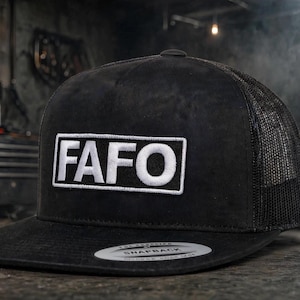 May include: A black snapback cap featuring a rectangular white patch on the front with the black text FAFO printed in a bold sans-serif font. The hat has a flat brim and a mesh back panel, with a circular sticker on the brim surface.