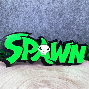 Puede incluir: Un letrero tridimensional que muestra la palabra SPAWN en letras de color verde neón brillante con un borde negro grueso. Un pequeño icono de calavera blanca con cuencas negras y boca dentada está situado en el centro de la letra A.