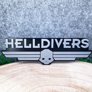 May include: A black and silver sign featuring the word HELLDIVERS in bold, blocky capital letters. Below the text is a stylised emblem consisting of a central skull icon flanked by horizontal wings, all rendered in a matte finish.