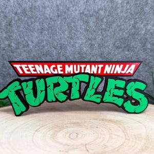 Op de afbeelding: Een driedimensionaal bord met het Teenage Mutant Ninja Turtles logo. Het bovenste gedeelte toont de woorden Teenage Mutant Ninja Turtles in witte tekst op een rode achtergrond, terwijl het woord Turtles in grote, felgroene letters staat.
