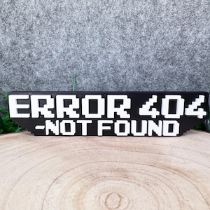 Könnte beinhalten: Ein schwarzes, rechteckiges Schild mit weißen, pixeligen Blockbuchstaben, die den Text ERROR 404 -NOT FOUND bilden. Die erhabene Schriftart verleiht dem Objekt eine digitale Retro-Optik mit einem klaren, kontrastreichen zweifarbigen Design.