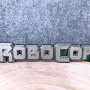 May include: A three-dimensional sign featuring the word ROBOCOP in a metallic silver finish with a blocky, industrial font style. The letters have a textured surface and are connected in a single piece, displaying a bold, sharp design.