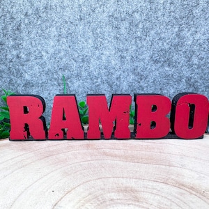 Könnte beinhalten: Ein freistehendes Schild mit dem Wort RAMBO in fetten Blockbuchstaben. Die Buchstaben haben eine leuchtend rote Vorderseite mit einer abgenutzten, strukturierten Oberfläche sowie eine schwarze Basis und Seitenkanten für einen Kontrast.