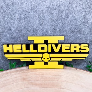 Pode incluir: Uma placa tridimensional com o texto amarelo HELLDIVERS II sobre um fundo preto. O design inclui um emblema de caveira amarela estilizada centralizado abaixo do texto, ladeado por asas horizontais, tudo montado sobre uma base preta.