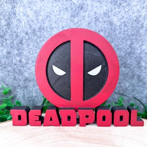 Może przedstawiać: Okrągły czerwono-czarny znak z logo Deadpoola z białymi kształtami oczu, umieszczony nad słowem DEADPOOL zapisanym pogrubionymi czerwonymi literami blokowymi. Znak ma teksturowane wykończenie i jest prezentowany jako samodzielna dekoracja.