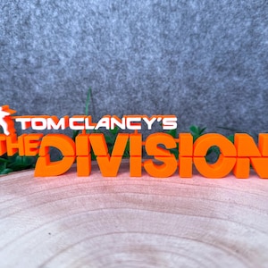 Könnte beinhalten: Ein leuchtend orangefarbenes 3D-gedrucktes Schild mit dem Schriftzug Tom Clancy's The Division in einer fetten, blockartigen Schriftart. Das Logo enthält eine kleine weiße Silhouette einer Person auf der linken Seite des Textes, alles im Querformat.