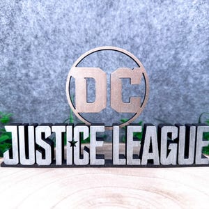 Könnte beinhalten: Ein dreidimensionales Schild mit dem DC-Logo in metallischer Bronzeoptik über dem Schriftzug JUSTICE LEAGUE in silbernen Blockbuchstaben. Die gesamte Struktur ist auf einem schwarzen Sockel mit einem kleinen Sternendetail montiert.