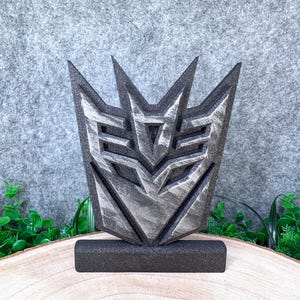 Könnte beinhalten: Eine dreidimensionale Decepticon-Emblem-Skulptur mit dunklem Metallic-Finish und silbernen Akzenten. Das schildförmige Objekt hat scharfe, kantige Spitzen und ein vertieftes geometrisches Design, montiert auf einem soliden rechteckigen Sockel.