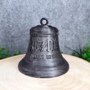 May include: A dark grey metallic bell featuring a raised AC/DC logo with a lightning bolt in the centre and the text HELL'S BELL embossed below. The top of the bell includes a small circular loop for hanging and text reading LOUGHBOROUGH.