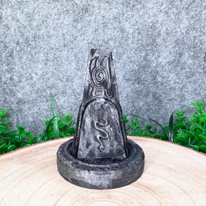 Peut inclure: Une sculpture en forme d'obélisque gris foncé, à l'aspect pierre vieillie, présentant un motif en spirale sculpté au sommet et un serpent au centre. L'obélisque est monté sur une base circulaire texturée avec une finition patinée assortie.