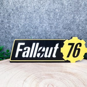 May include: A black rectangular sign featuring the white text Fallout 76 with a lightning bolt icon inside the letter o. To the right of the text is a yellow gear-shaped emblem with the number 76 in black. The sign has a thin yellow border.