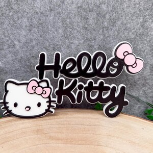 Op de afbeelding: Een Hello Kitty embleem met een wit kattengezicht met zwarte omlijning, een roze strik en de zwarte tekst Hello Kitty in een cursief lettertype met een bijpassende roze strik aan de rechterkant van de letters, alles met een witte rand.