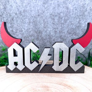 Op de afbeelding: Een decoratief bord met de zilverkleurige tekst AC/DC en een centraal bliksemschichtsymbool. De letters zijn gemonteerd op een zwarte basis en worden geflankeerd door twee rode duivelshoorns die omhoog steken vanaf de buitenste letters.