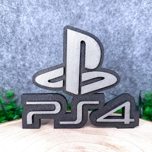 May include: A three-dimensional PlayStation 4 logo sign featuring a black base with a silver-colored top layer. The design includes the iconic PlayStation symbol positioned above the text PS4, rendered in a bold, blocky font style.