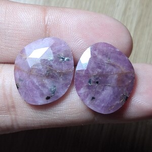 Op de afbeelding: Twee ovaalvormige, gefacetteerde roze edelstenen met zichtbare natuurlijke insluitingen en donkere mineraalvlekken. De stenen hebben een gepolijst, reflecterend oppervlak met een doorschijnende kwaliteit, met variaties in licht- en donkerroze tinten door het materiaal.