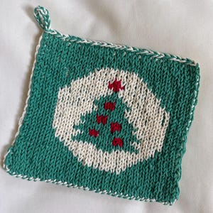Può includere: Un sottopentola quadrato lavorato a maglia con un motivo a forma di albero di Natale. Il sottopentola è principalmente verde acqua con una forma di albero bianca al centro, adornata da accenti rossi. Ha un bordo bianco e verde e un anello per appenderlo.