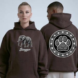 May include: A dark brown hooded sweatshirt featuring a graphic of an elephant with the text "Swagger" on the front. The back of the hoodie displays a large circular emblem containing a paw print design surrounded by a border of white stars.