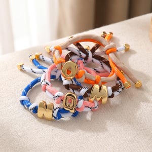 May include: A collection of braided cord bracelets in blue, pink, orange, and brown, each featuring a polished gold-tone metal letter charm. The cords are woven with white accents and finished with small gold-tone metal beads at the ends.