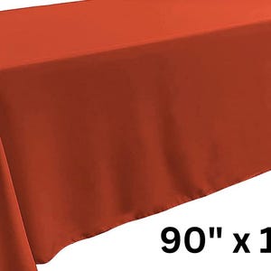 May include: A solid burnt orange rectangular tablecloth draped over a table, featuring a smooth, matte fabric texture. The dimensions 90" x 132" are clearly printed in black text in the lower right corner of the image against a white background.
