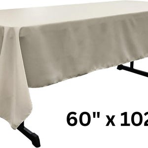 May include: A rectangular beige tablecloth draped over a black folding table. The fabric has a smooth, solid-colored finish and hangs down on all sides. The text 60" x 102" is printed in black in the lower right corner, indicating the dimensions.