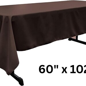 May include: A solid dark brown rectangular tablecloth draped over a black folding table. The fabric has a smooth, matte finish and hangs evenly on all sides. The dimensions 60" x 102" are printed in black text in the lower right corner of the image.