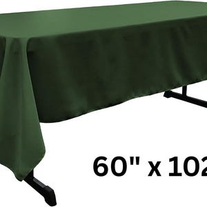 May include: A solid dark green rectangular tablecloth draped over a black folding table. The fabric has a smooth, matte finish and hangs down on all sides. The text 60" x 102" is printed in black in the lower right area of the image.