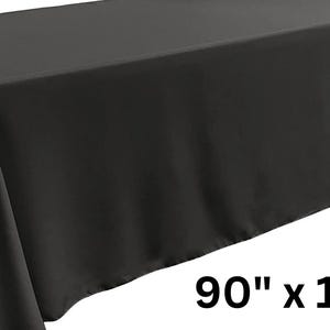 May include: A solid black rectangular tablecloth draped over a table, featuring a smooth, matte fabric texture. The text 90" x 132" is printed in the lower right corner, indicating the dimensions of this rectangular table covering.