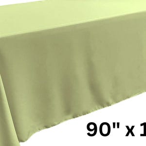 May include: A rectangular tablecloth in a solid light lime green color. The fabric drapes over the edges of a table, creating smooth folds. The text 90" x 132" is printed in black in the lower right corner, indicating the dimensions of the item.