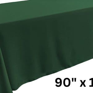 May include: A solid dark green rectangular tablecloth draped over a table, featuring a smooth, matte fabric texture. The dimensions 90" x 132" are printed in black text in the lower right corner of the image, indicating the size of the cloth.
