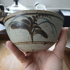 May include: A speckled ceramic bowl with a light beige base color, featuring a hand-painted dark brown botanical leaf design and thin blue arched lines. The bowl has a horizontal brown band near the rim and base, with a small stamped mark at the bottom.