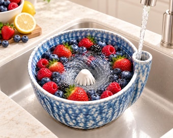 May include: A blue and white speckled ceramic berry colander bowl with a central white cone and a side spout. The bowl is filled with fresh strawberries, blueberries, and blackberries being rinsed under a stream of water from a kitchen faucet.