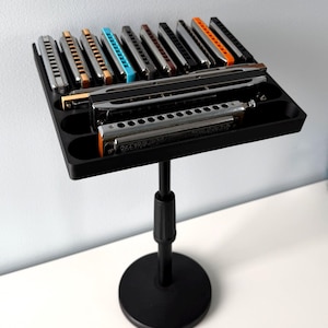 May include: A black adjustable stand featuring a rectangular tray with multiple slots holding a collection of harmonicas in various colors including silver, gold, blue, and orange. The stand has a circular base and a central vertical support pole.