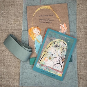 Könnte beinhalten: Ein beiges Buch mit dem Titel In Beziehung setzen mit goldenem Bogen und Figur, eine türkisfarbene Karte Nummer 29 mit einer Strichzeichnung einer Person mit Baby und ein salbeigrünes Gummiband auf einem hellblauen Filzblatt. Der Buchtitel lautet Impulse und Fragen für Bewusstheit, Wachstum und innere Wandlung.